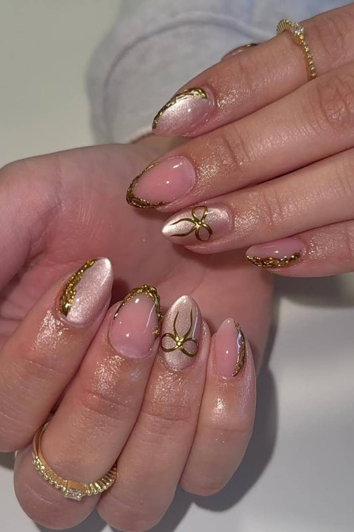 Close-up of hands with almond-shaped nails painted in shimmery nude polish, decorated with gold accents and fine gold line designs.