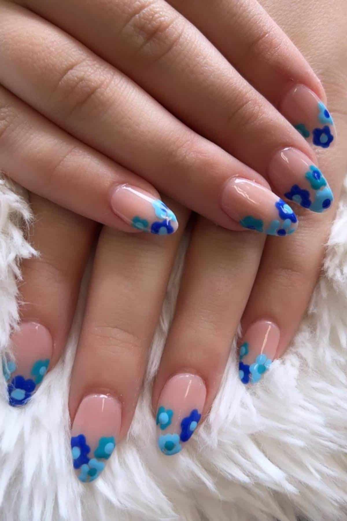 Close-up of hands with almond-shaped nails featuring nude polish and blue floral nail art tips, resting on a white furry surface.