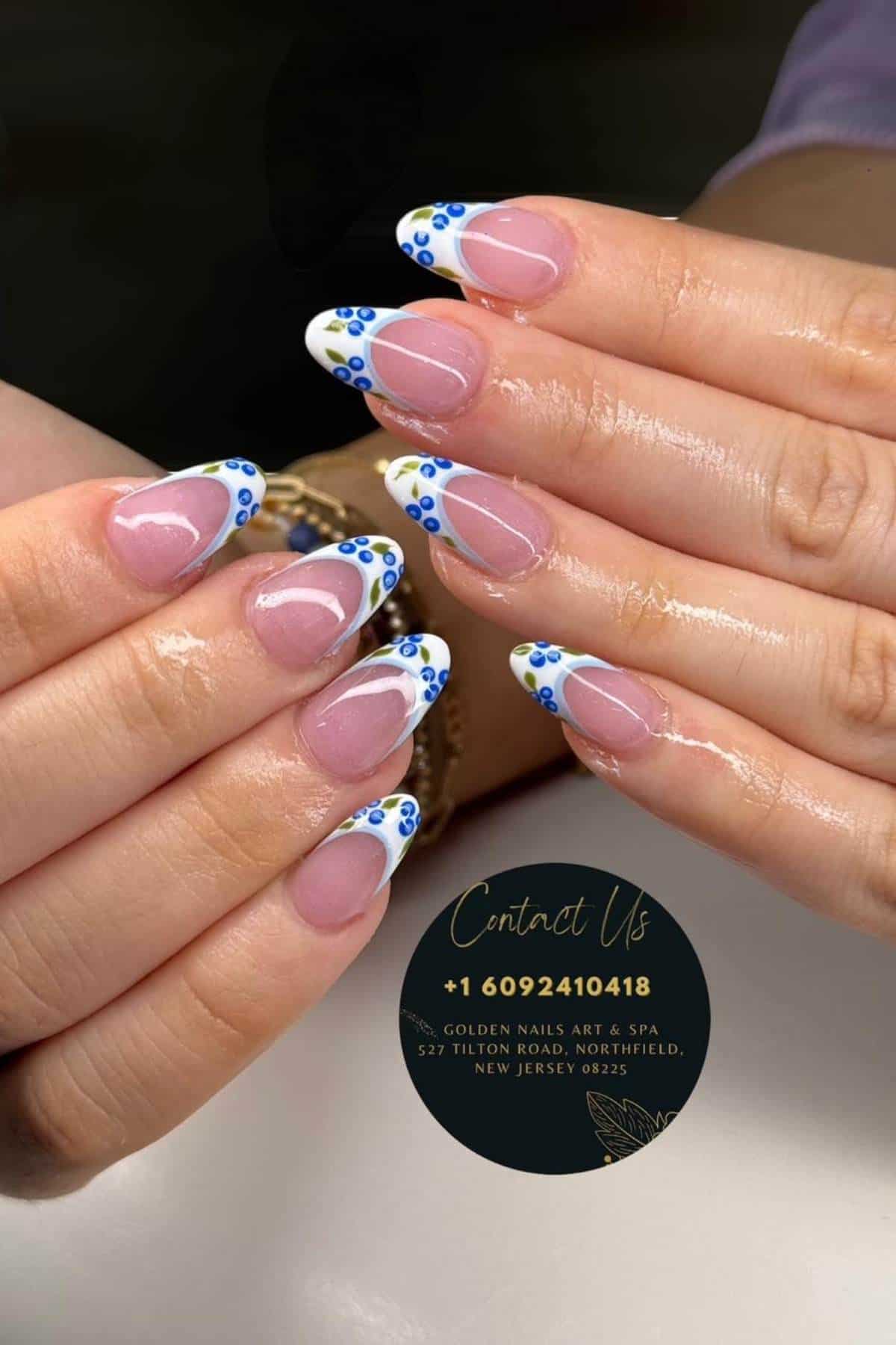 A close-up of hands with almond-shaped nails featuring French tips decorated with small blue and white floral designs. A contact card for Golden Nails Art & Spa is also visible.