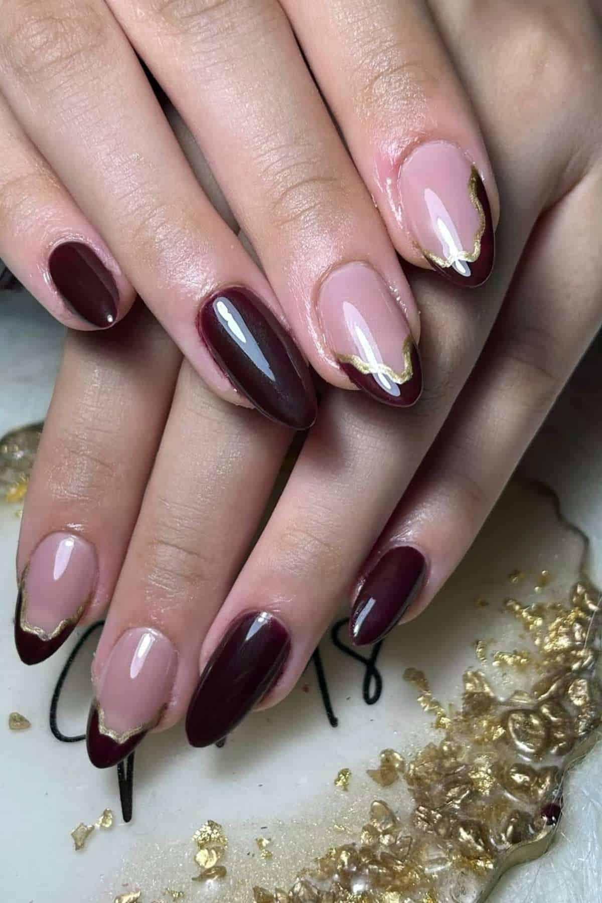 Hands with almond-shaped nails featuring a mix of maroon polish and nude polish with gold foil accents on the tips, displayed over a white and gold decorative surface.