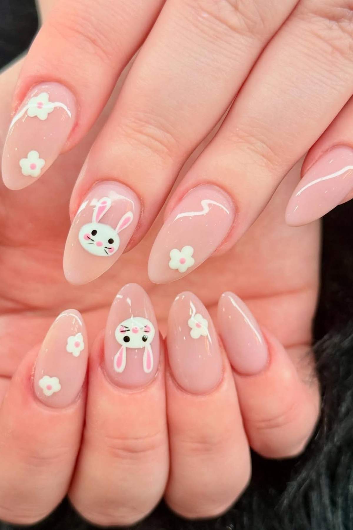 Hands with long, almond-shaped light pink nails featuring white flower designs and two accent nails with white cartoon bunny faces.