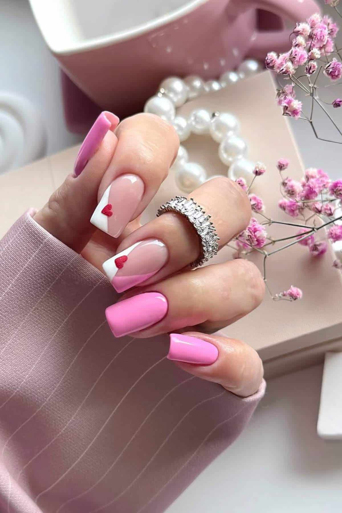 A hand with long pink and white manicured nails, two featuring small red heart designs, holding a pink cup, with pearls and pink flowers in the background.