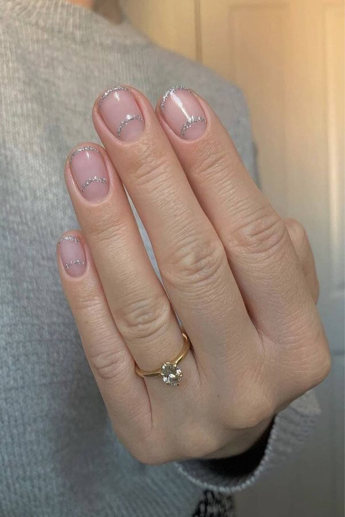 A hand with a gold ring and clear nails featuring thin, silver glitter tips; person is wearing a light gray sweater.