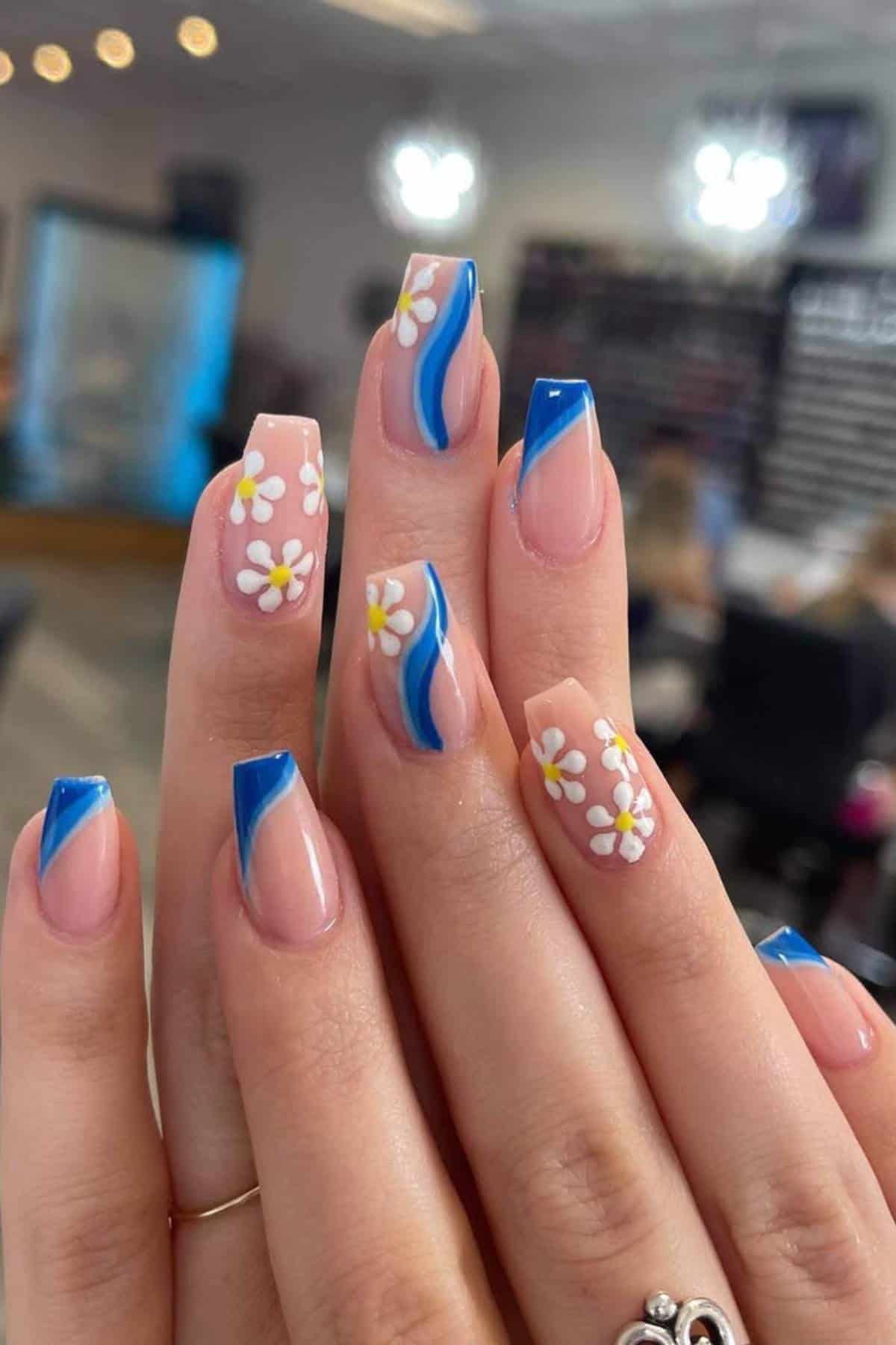 A hand with square-shaped nails featuring blue French tips, blue swirl accents, and white daisy flower designs on a nude base.