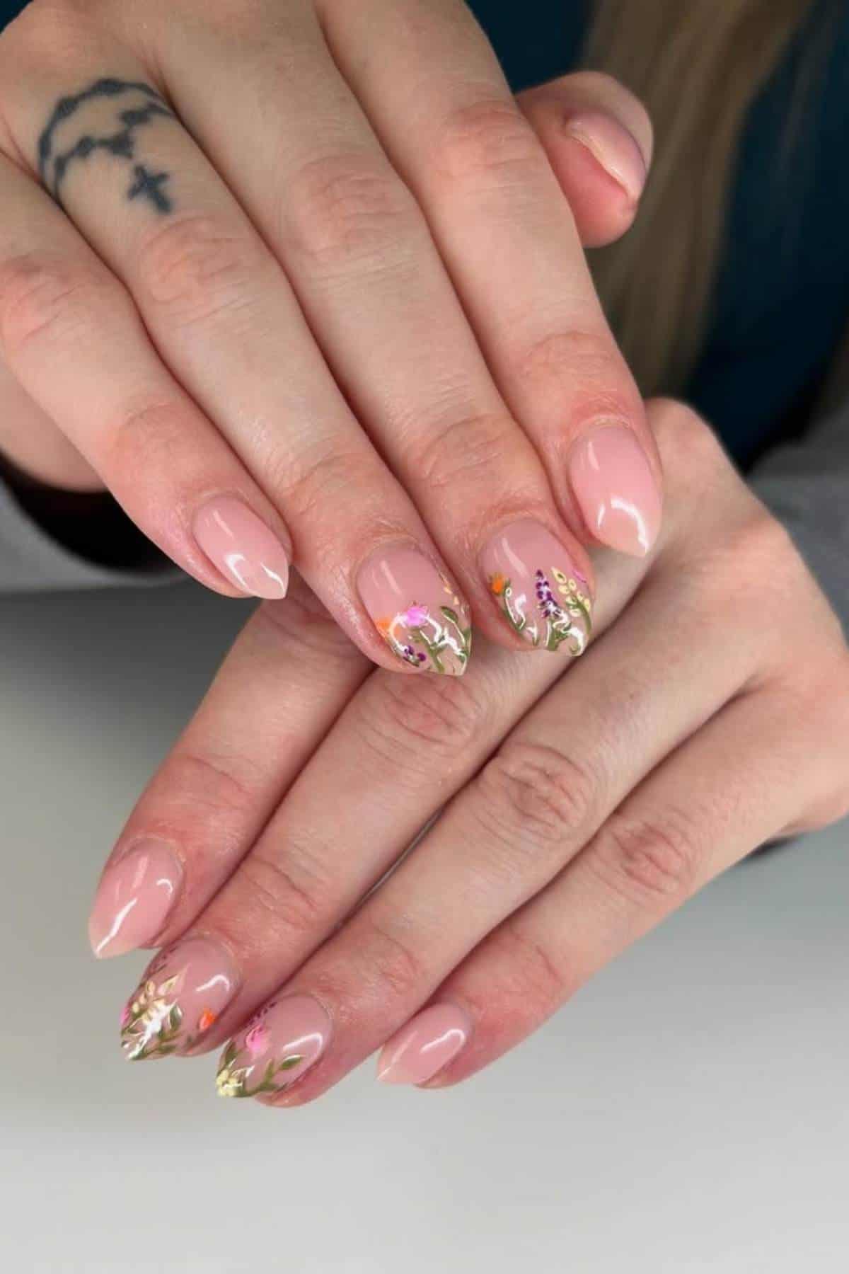 A hand with nude almond-shaped nails featuring delicate floral nail art; a tattoo of three small birds is visible on one finger.