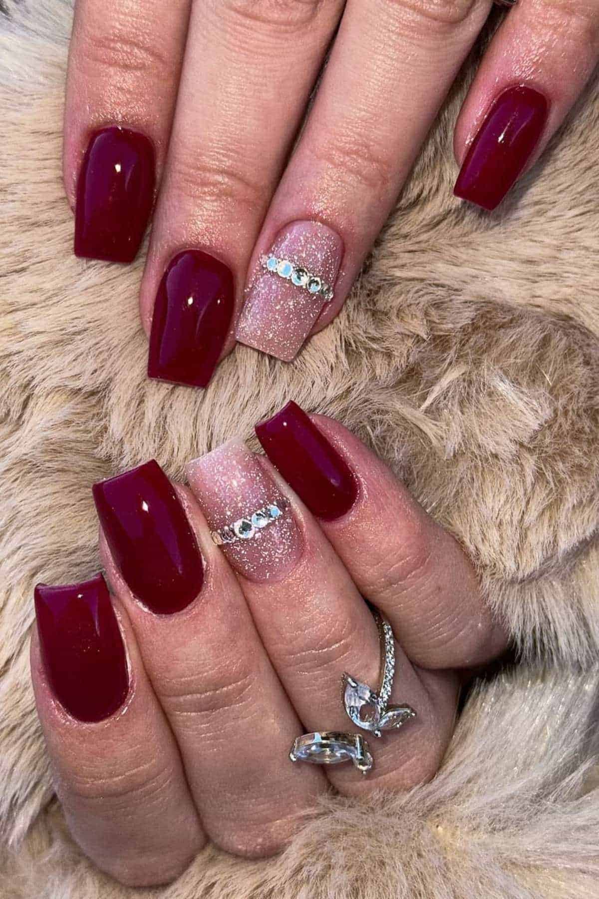 Hands with square-shaped burgundy nails, one accent nail on each hand in glittery nude with rhinestone decoration, resting on a soft beige fabric.