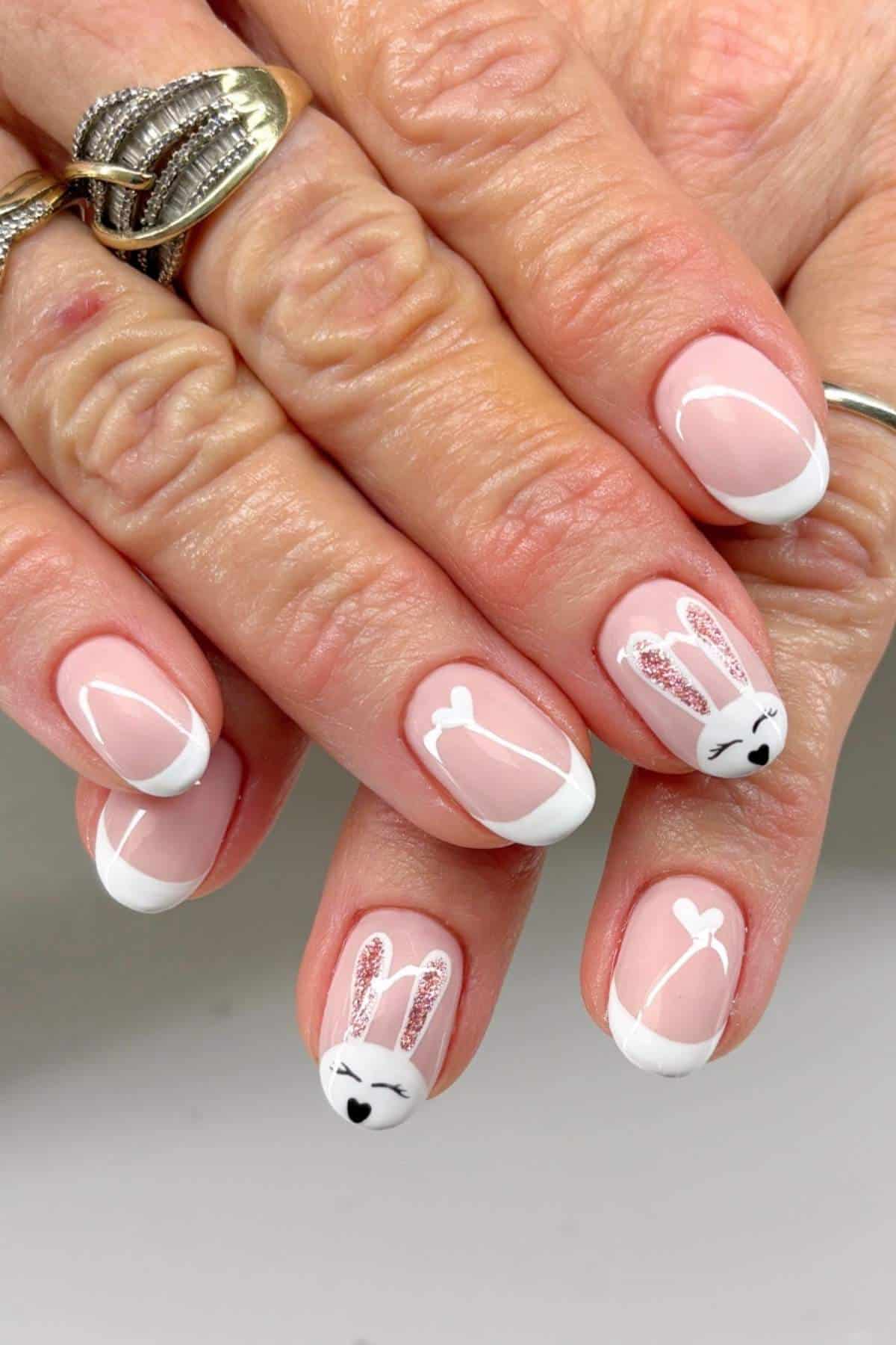 Hands with a French manicure featuring white tips and accent nails decorated with white bunny faces and ears, along with silver glitter details.