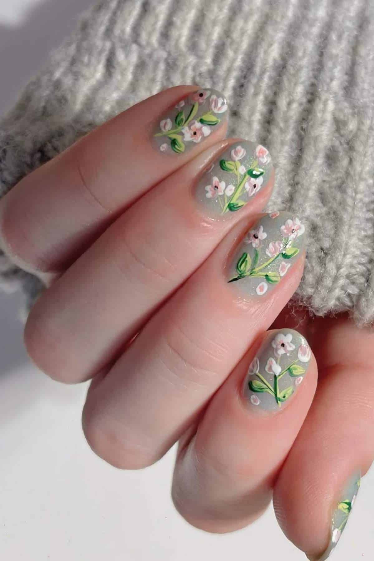 A close-up of a hand with light grey nail polish featuring small white floral designs and green leaves, against a grey knitted sleeve.