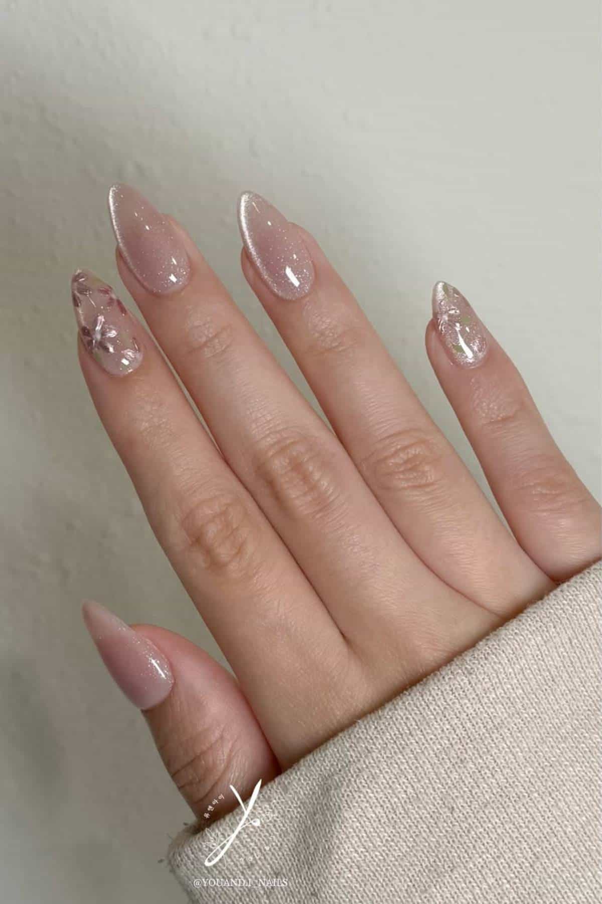 A hand with almond-shaped nails painted in light pink polish, some with glitter and floral decorations, resting on a light surface.