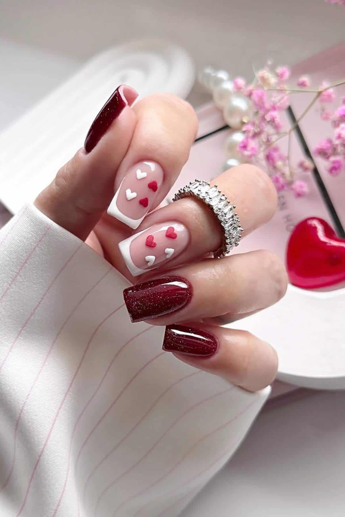 A hand with manicured nails featuring dark red polish and white accent nails with pink and red heart designs, holding a pearl ring, with decorative items in the background.