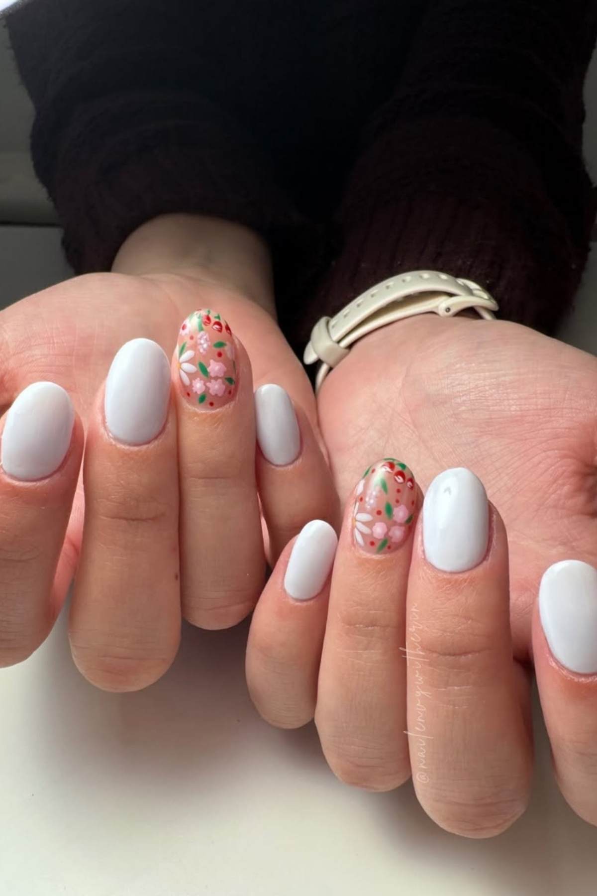 Hands with short, oval-shaped nails painted white; the ring fingers feature floral designs in pink, red, and green.