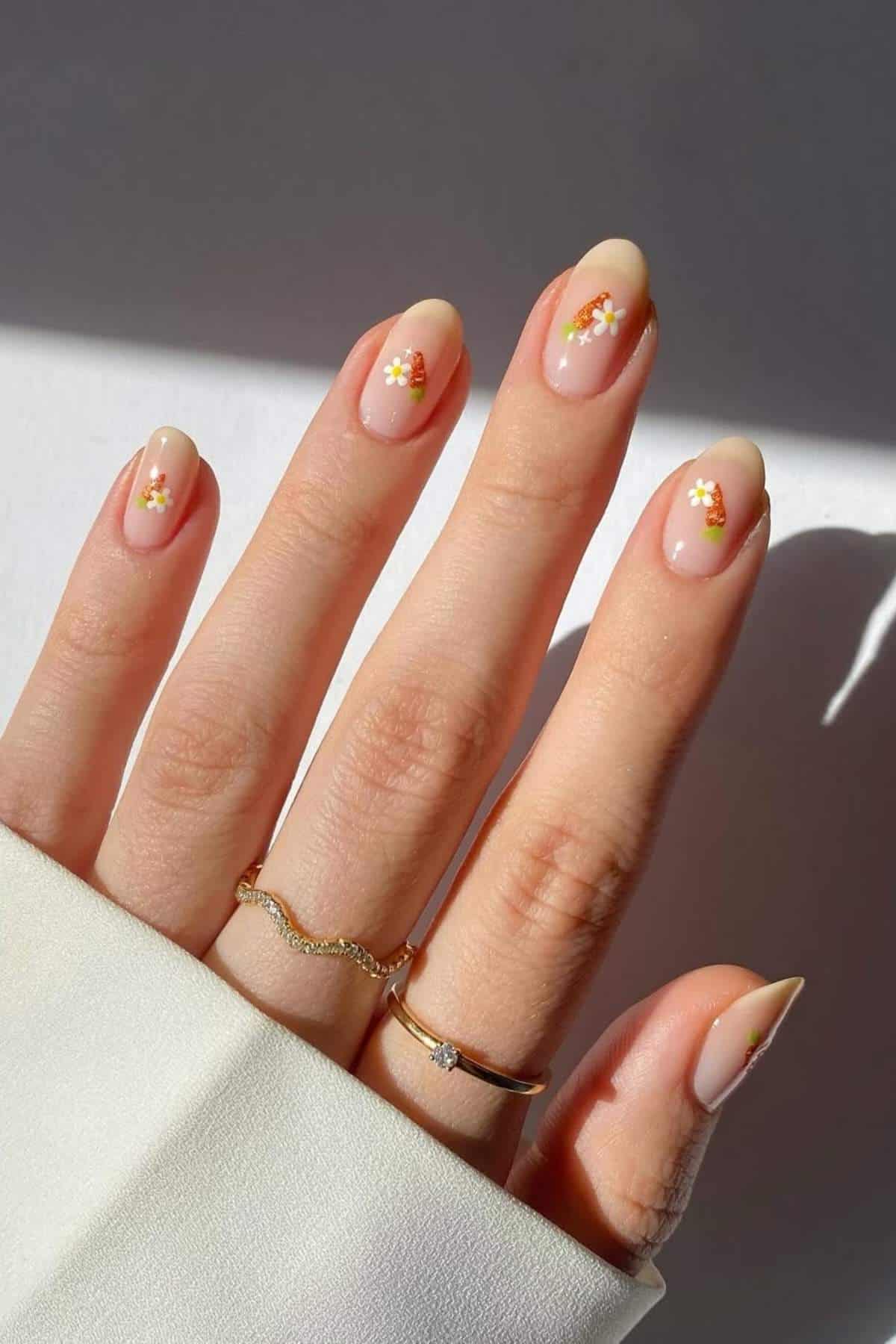 A hand with almond-shaped nails features a nude base and small white daisy designs on each nail. The person is wearing two gold rings and a white sleeve.