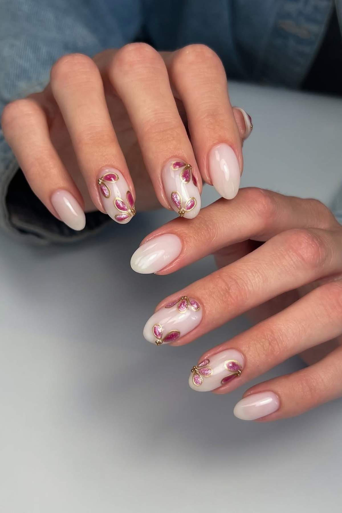 A person's hands with long, almond-shaped nails painted in a pale pink shade, featuring pink and gold floral nail art designs.