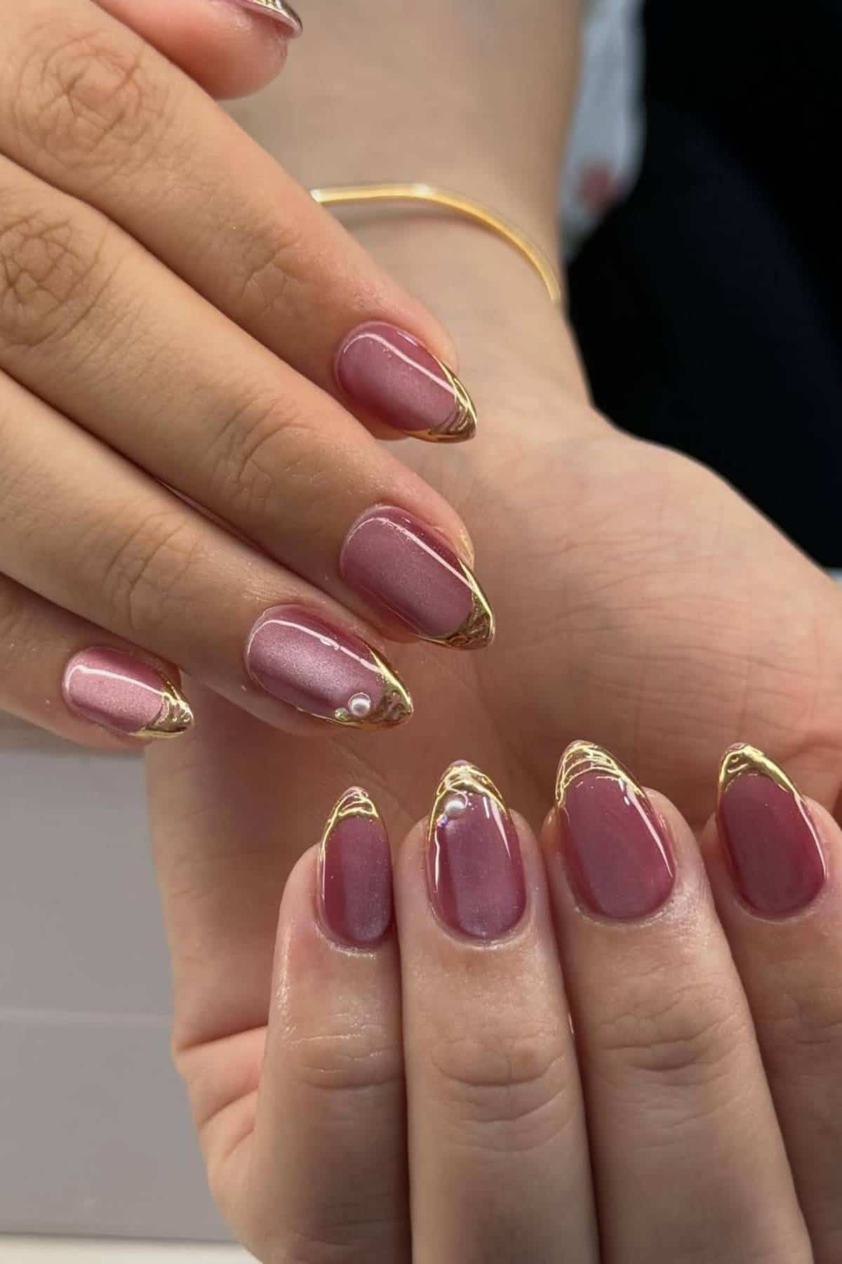 Close-up of hands with almond-shaped nails painted mauve and decorated with thin gold metallic lines on the tips.