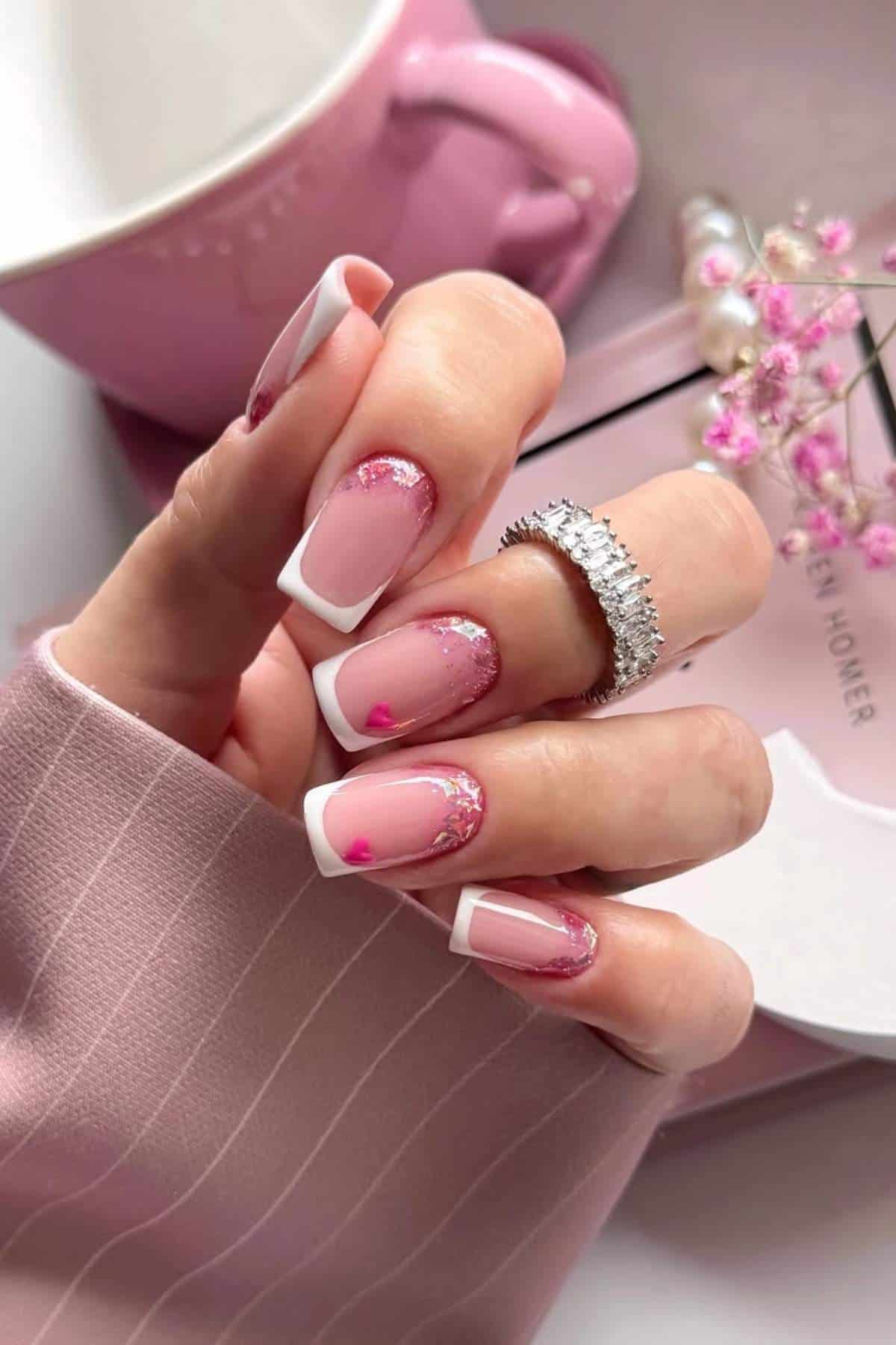 A hand with manicured nails featuring pink polish, white French tips, small heart accents, and glitter, holding a pink cup. The person wears a silver ring and a pink pinstripe sleeve.