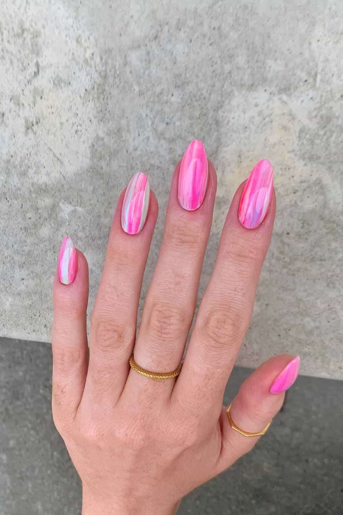 A hand with long, almond-shaped nails featuring a pink and white marbled design, wearing two thin gold rings on the index and ring fingers.