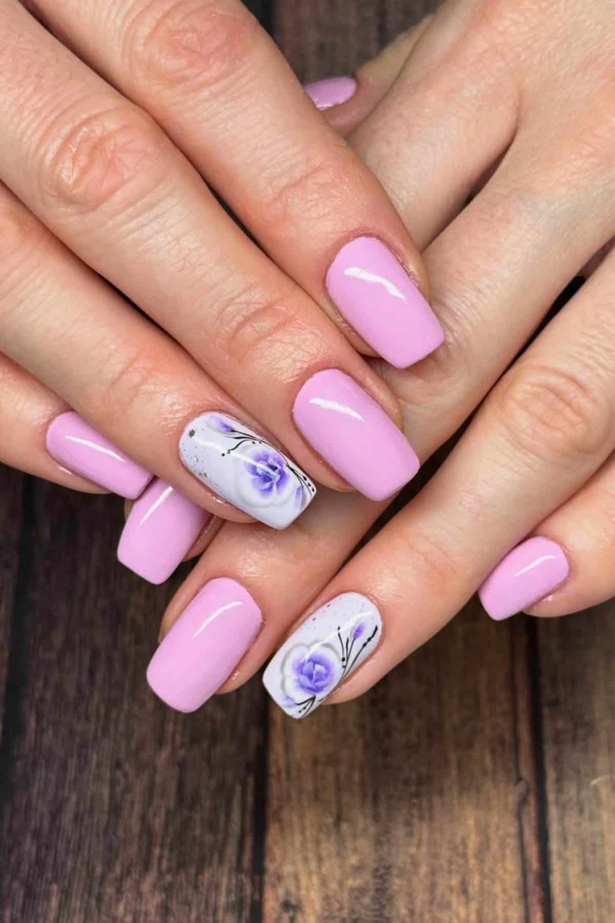 Hands with short, square-shaped nails painted light pink; ring fingernails feature a purple flower design with thin black accents on a white background.