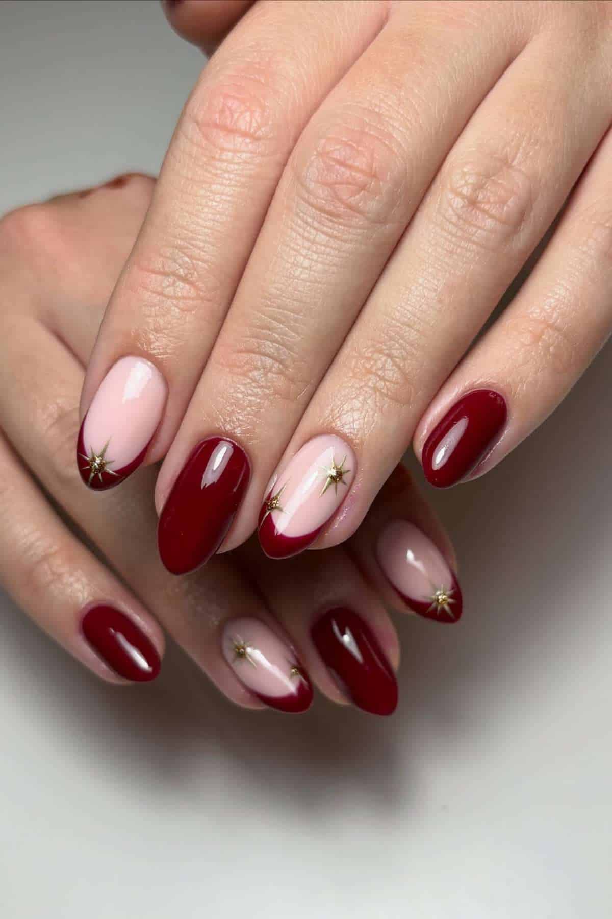 A hand with manicured nails featuring a mix of deep red, light pink, and red French tips with small gold star accents.
