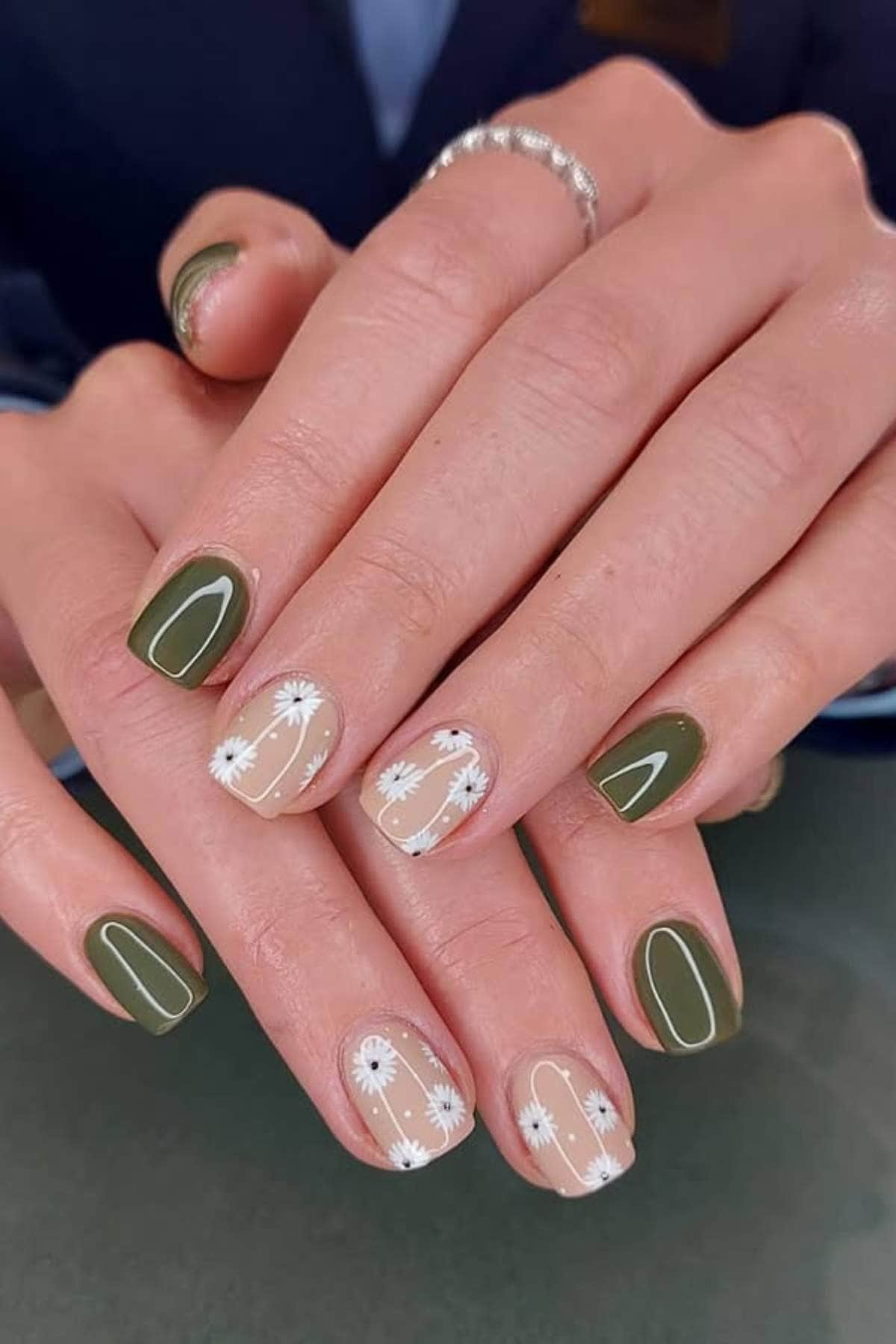 Hands with manicured nails; some nails are painted olive green, while others are nude with white floral and line designs.