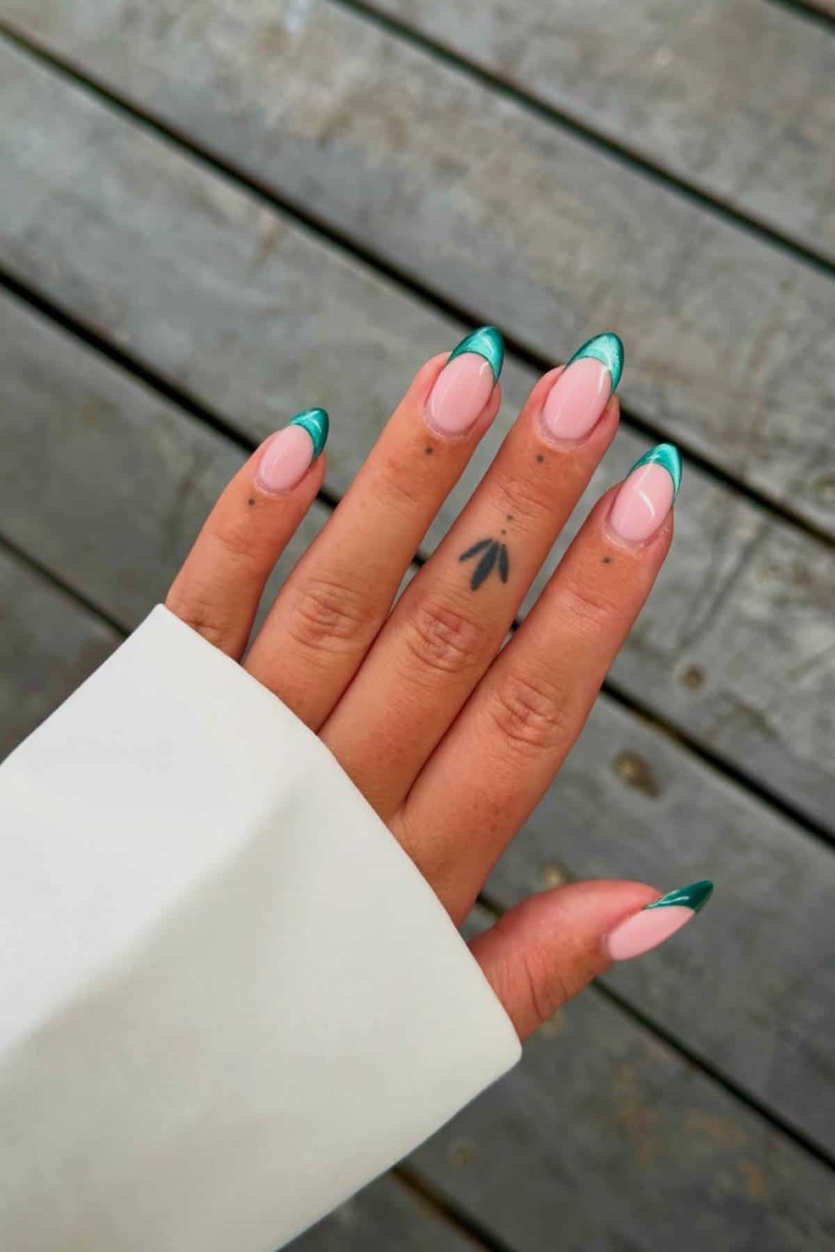 A hand with almond-shaped nails painted with a pink base and teal French tips; a small black tattoo is visible on the middle finger. The background is weathered wooden planks.