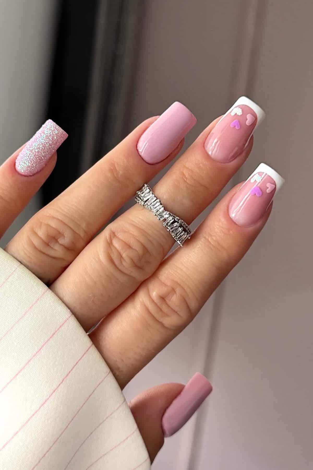 A hand with long, square-shaped nails painted pink; some nails have white tips, heart designs, and glitter accents. The person is wearing a silver ring.