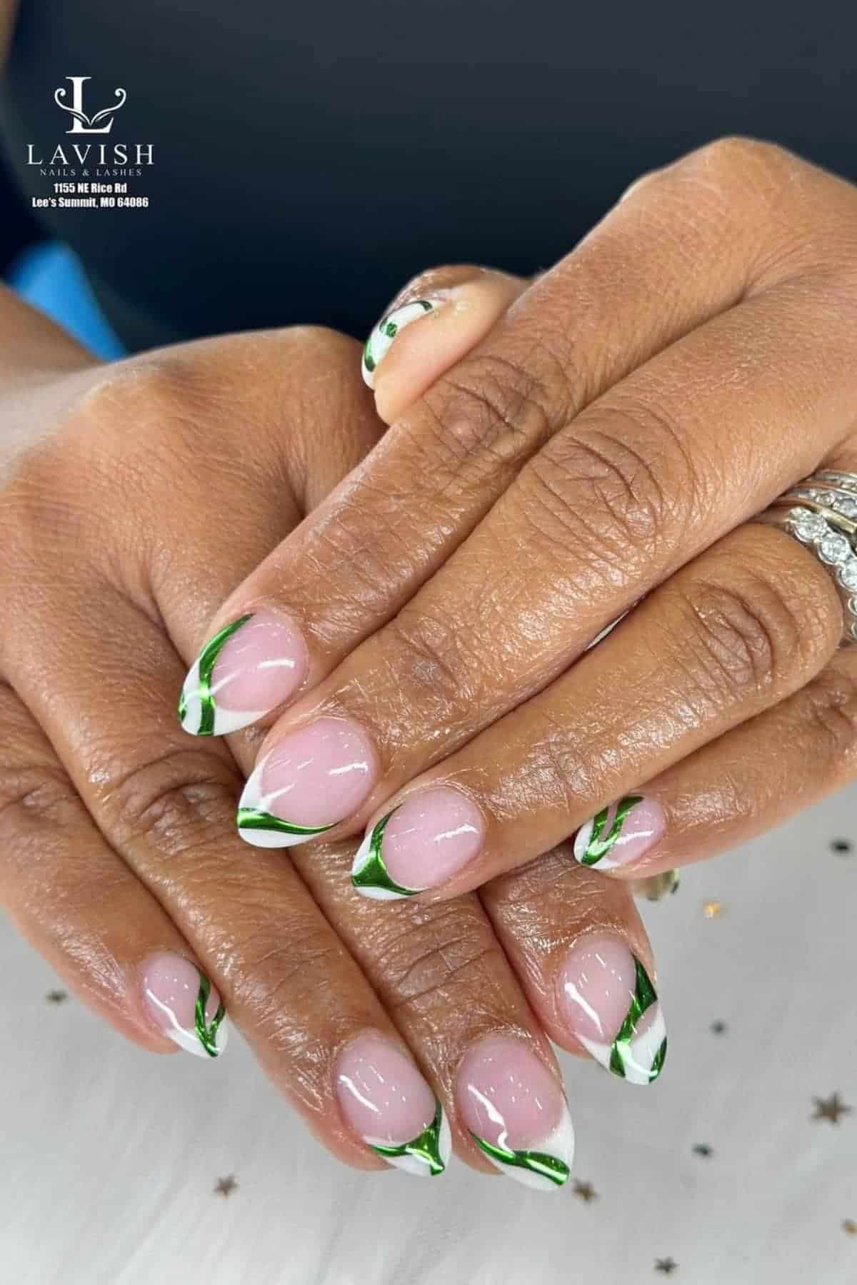 Hands displaying manicured nails with a clear base and green and white abstract designs on the tips, adorned with rings.