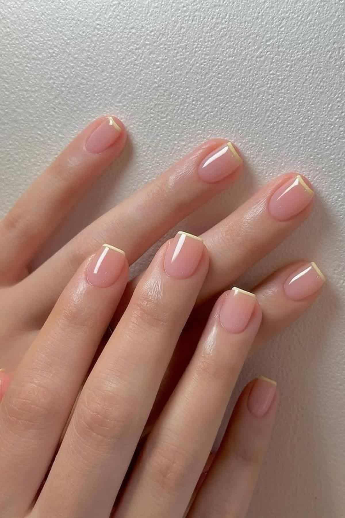 A hand with short, natural-shaped nails featuring a nude pink base and thin, pale yellow French tips, resting on a light textured surface.