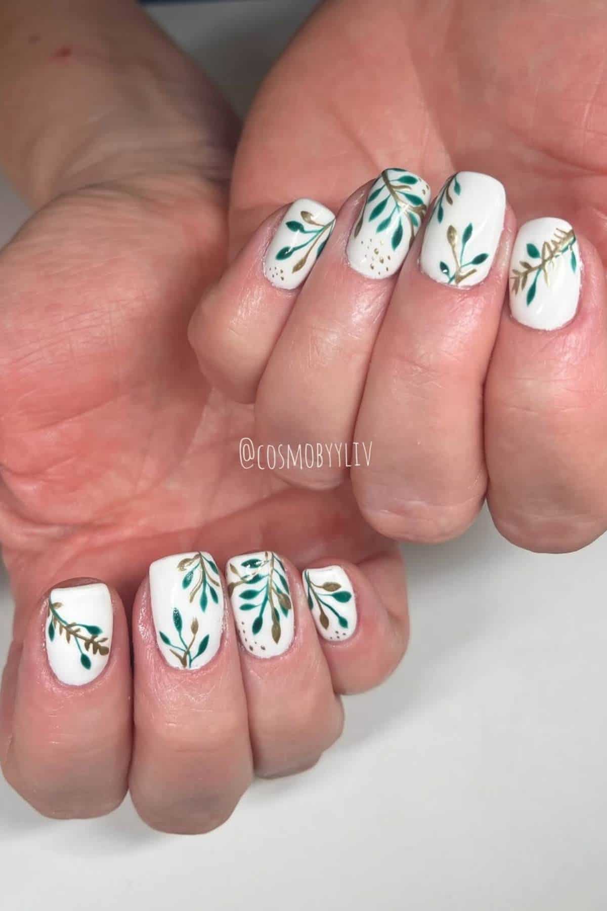 Hands with white nail polish featuring green and gold leafy branch designs on each nail.