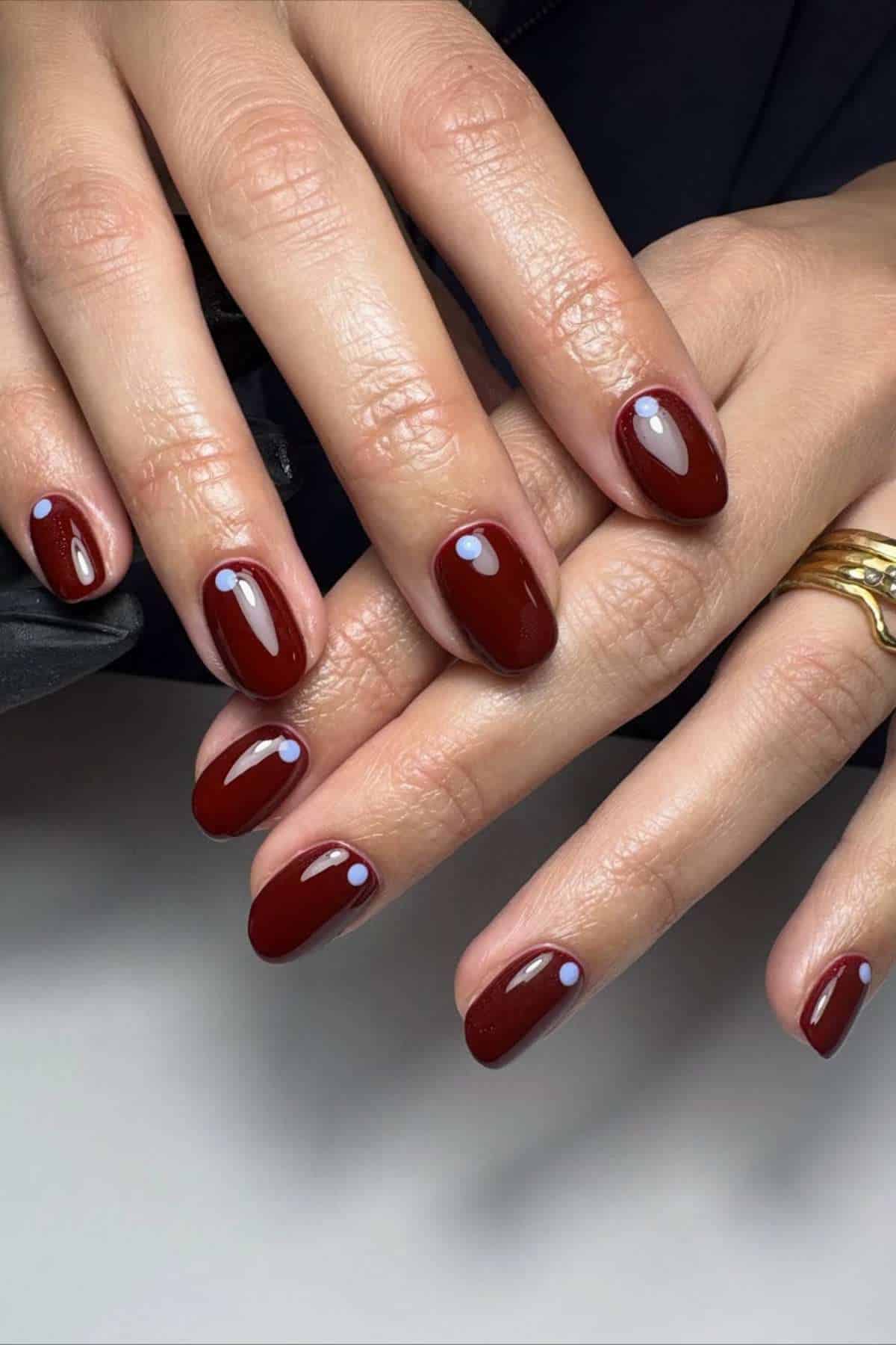 Hands with dark red almond-shaped nails, each featuring a small light lavender dot near the cuticle; fingers are adorned with gold rings.