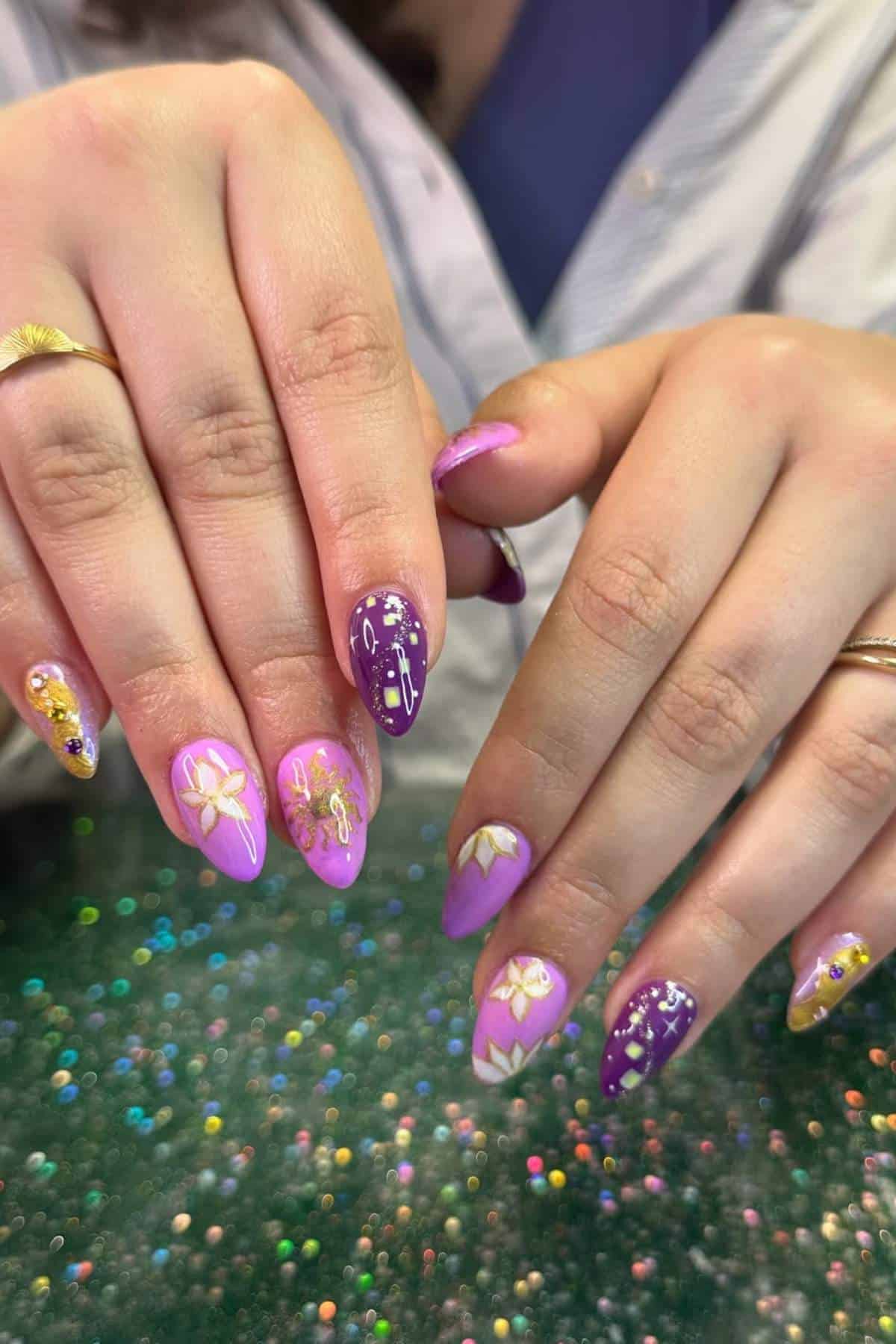 A person displays manicured nails with purple and pink polish, featuring gold floral and star designs, over a glittery green surface.