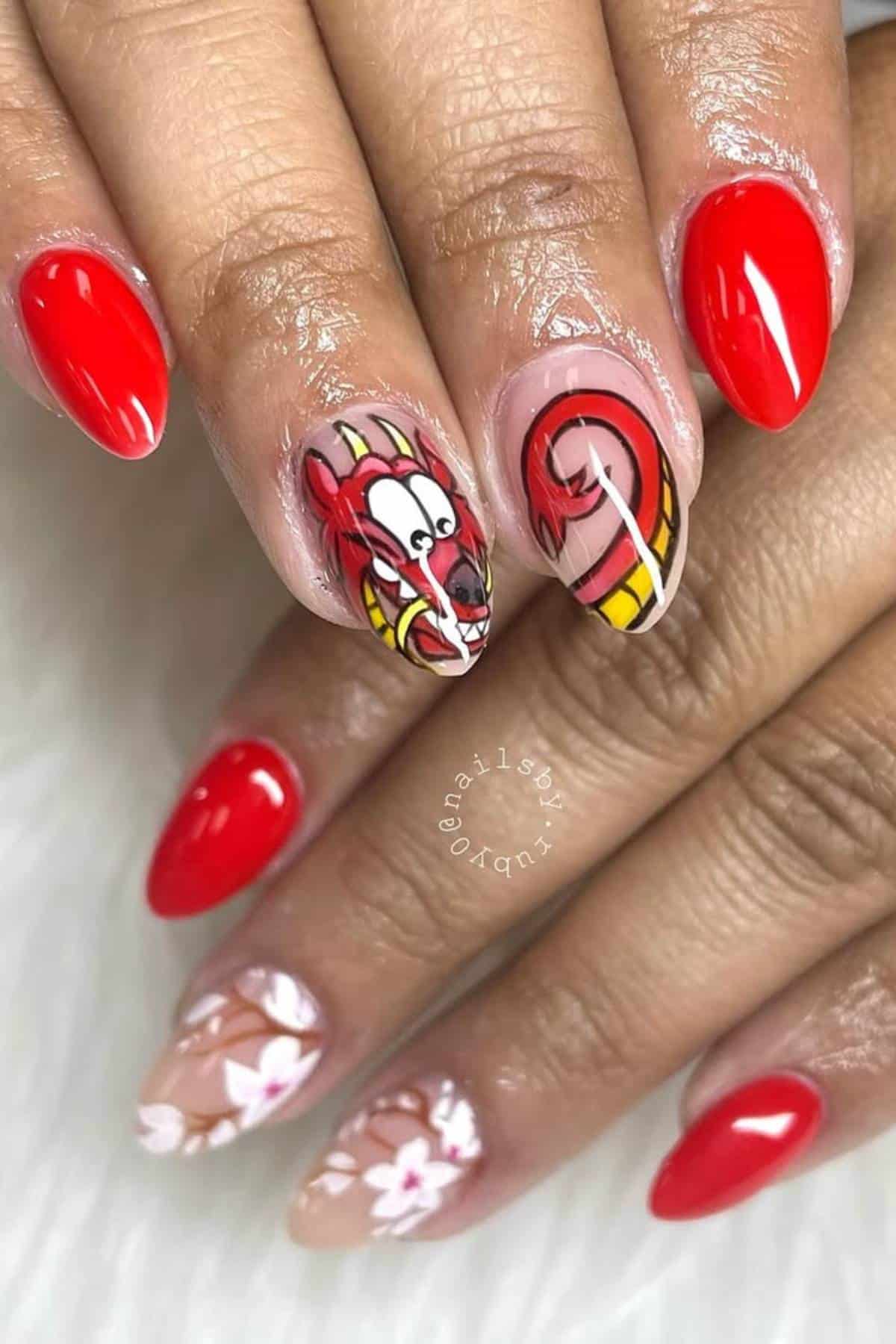 A hand with red, almond-shaped nails; two accent nails feature cartoon dragon art and floral designs on a nude base.