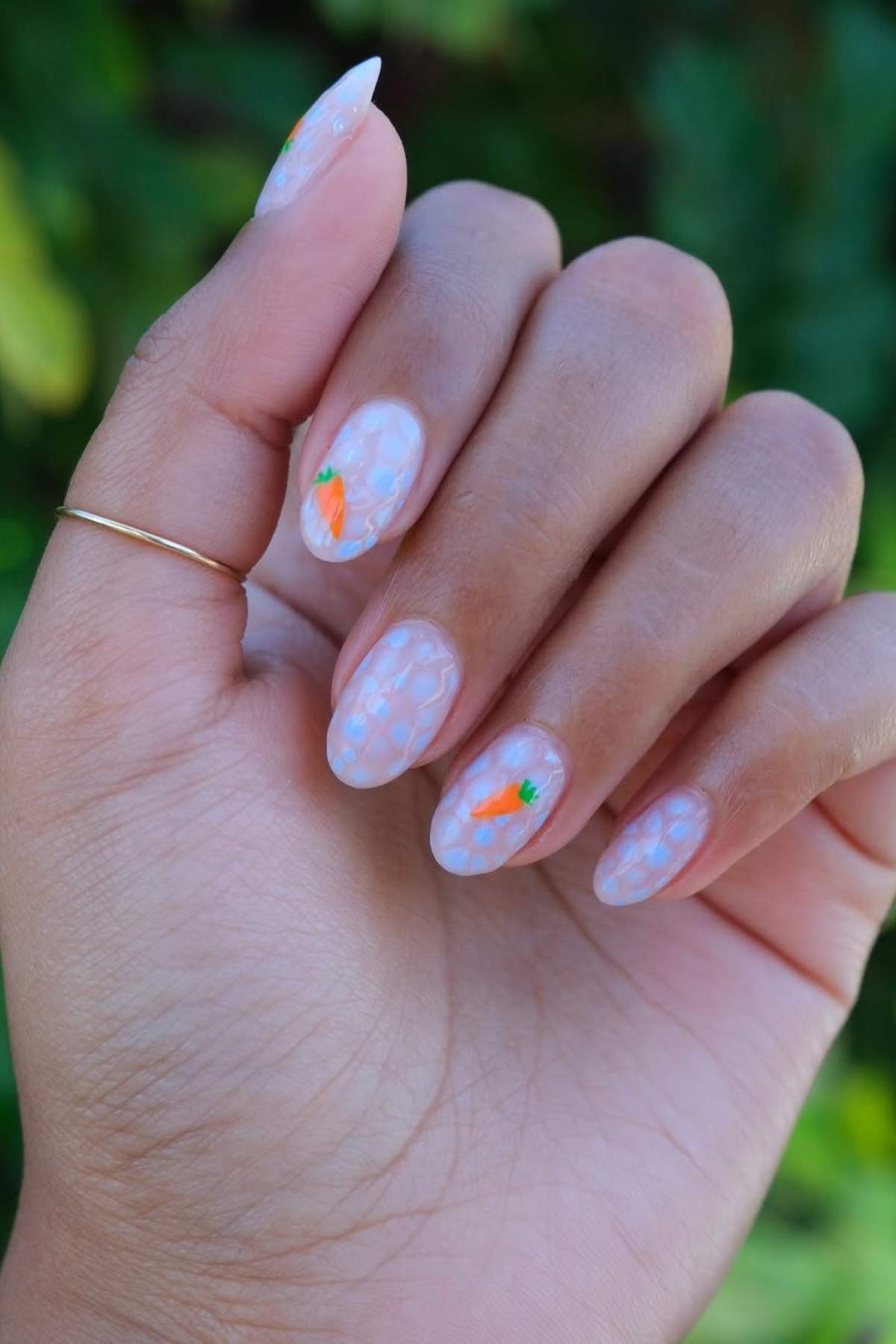 A hand with manicured nails featuring a light blue dot pattern and small painted carrots on two nails.