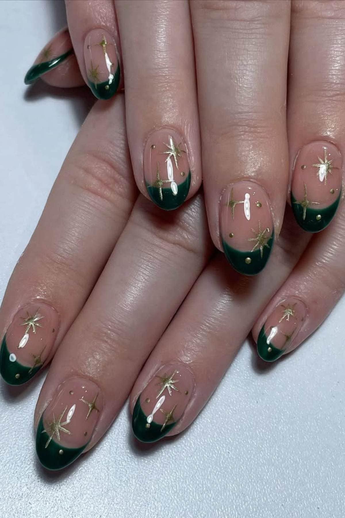 Hands with almond-shaped nails featuring dark green French tips and gold starburst designs on a natural base.