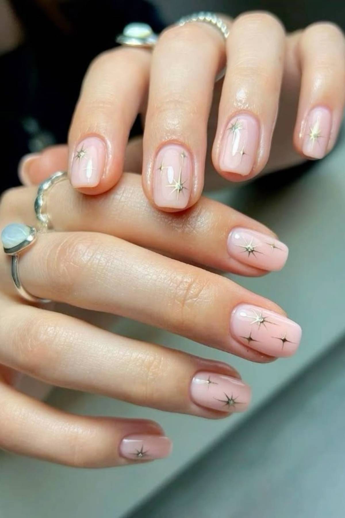 A hand with short, square-shaped nails painted in nude pink polish and decorated with small gold star designs.