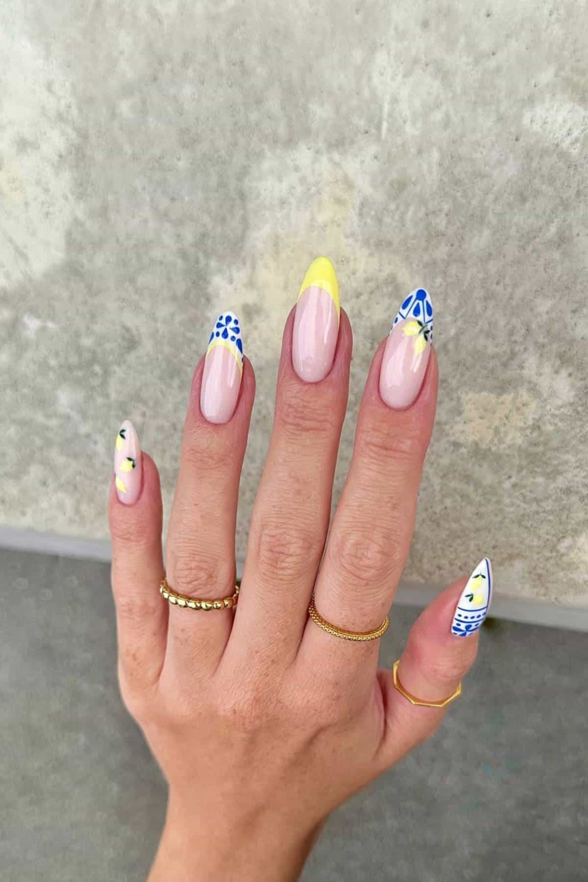 A hand with long, almond-shaped nails featuring yellow tips and blue floral designs, wearing gold rings, against a textured gray background.