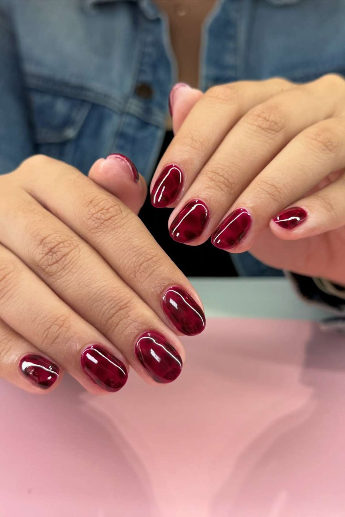 Close-up of hands with short, oval-shaped nails painted in glossy dark red polish against a light pink surface. The person is wearing a blue denim shirt.