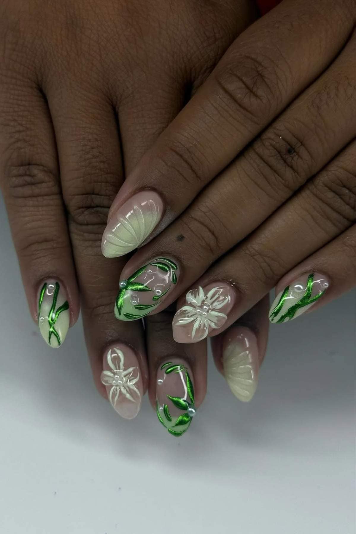 Close-up of hands with almond-shaped nails featuring white and green floral and leaf designs, some with small decorative pearls, on a natural beige base.