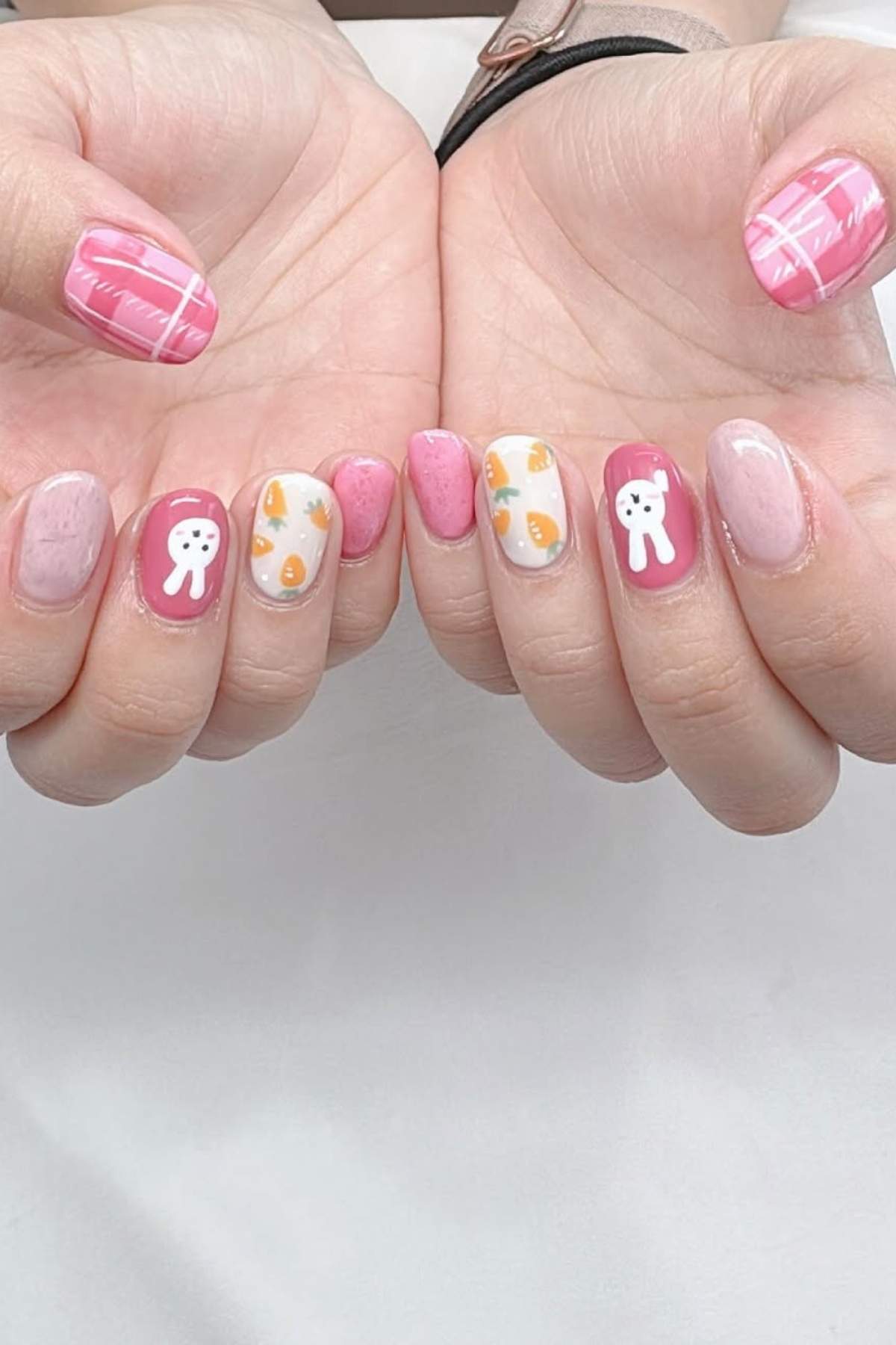 Hands with pink and white manicure featuring patterns of teeth, cheese slices, pink hearts, and plaid designs on short nails.