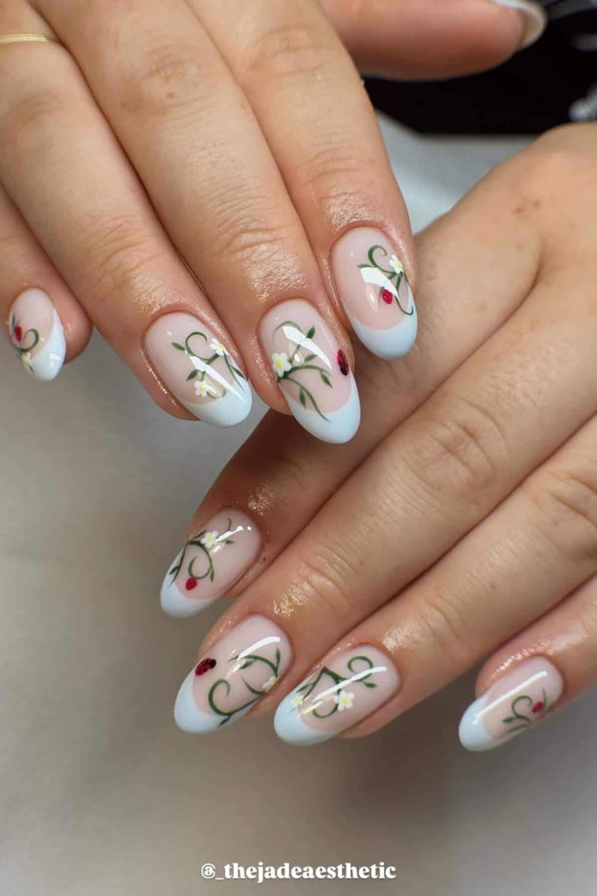 Hands with almond-shaped nails featuring a light blue French tip and floral vine designs with small red and white accents on a nude base.