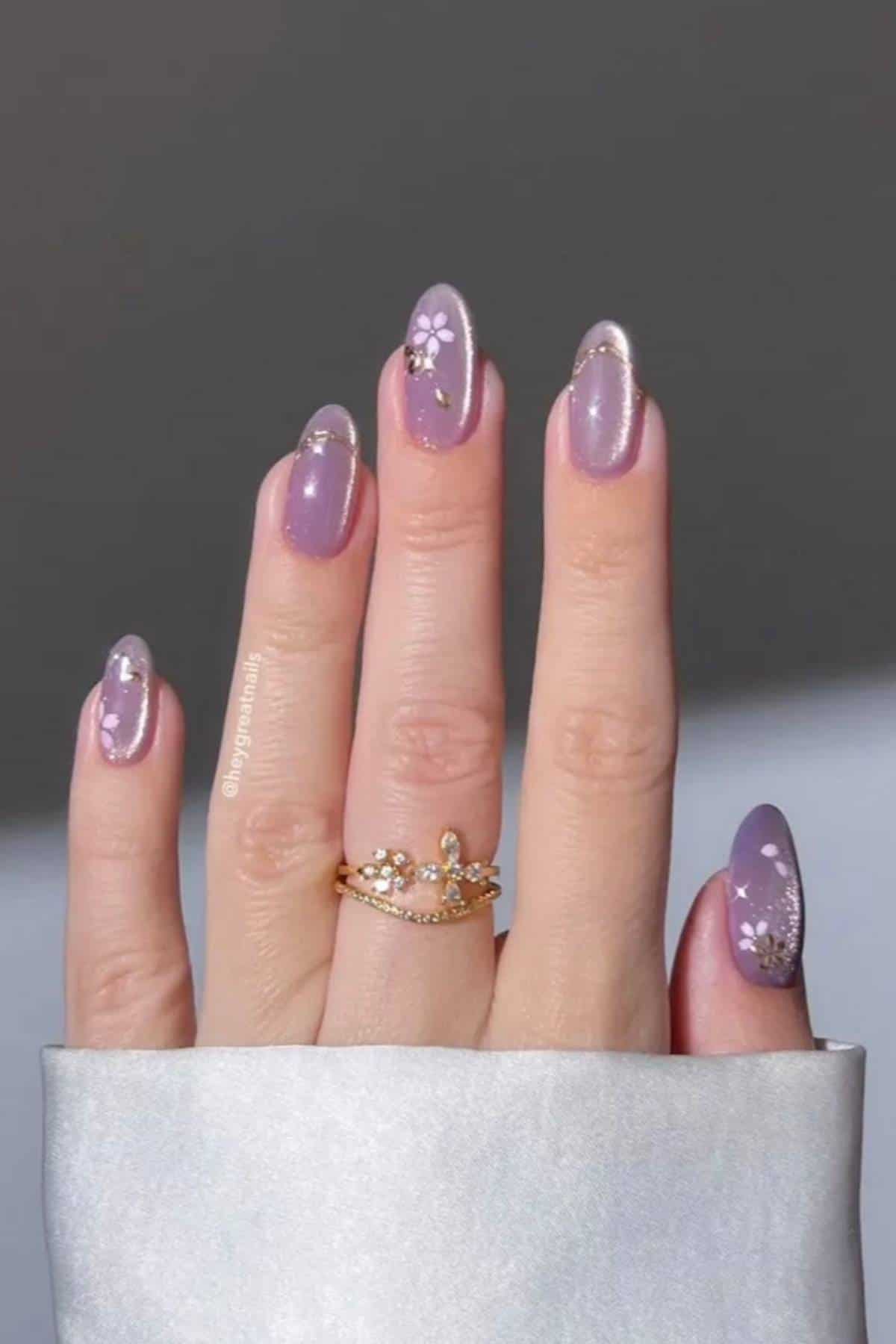 A hand with long, almond-shaped nails painted lavender, featuring white floral designs and small gemstones, wearing a gold crown-shaped ring.