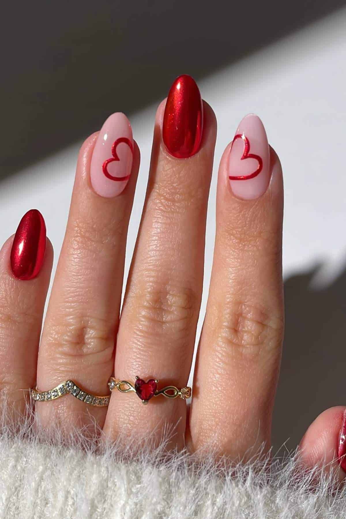 A hand with four fingernails painted red and pink; pink nails have outlined red heart designs. The person wears two gold rings, one with a red heart-shaped stone.