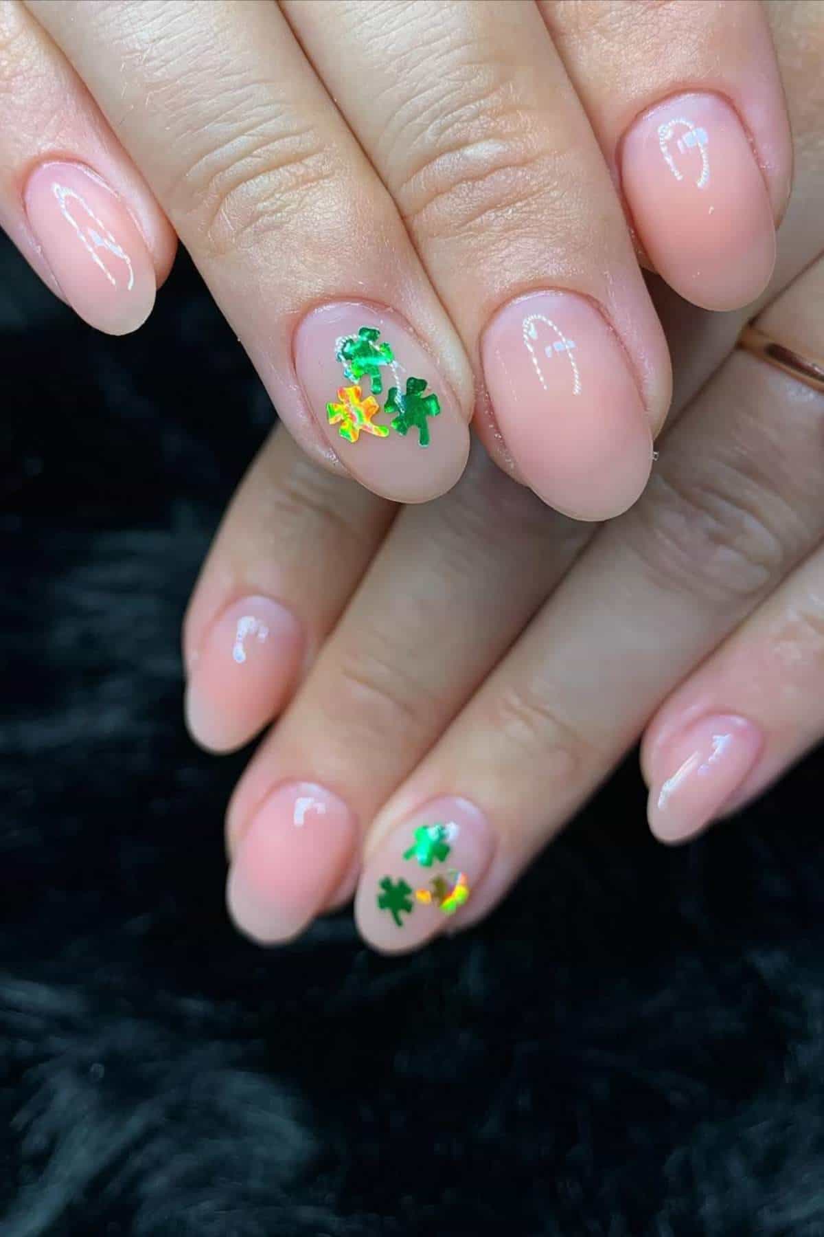 Close-up of hands with short oval nails painted in a nude color, featuring small green and yellow shamrock-shaped nail art on two accent nails.