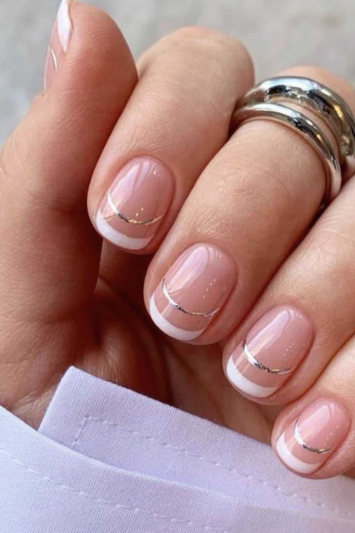 A hand with short, oval-shaped nails featuring a pale pink base, white French tips, and a thin silver line below each tip. The person is wearing two silver rings.