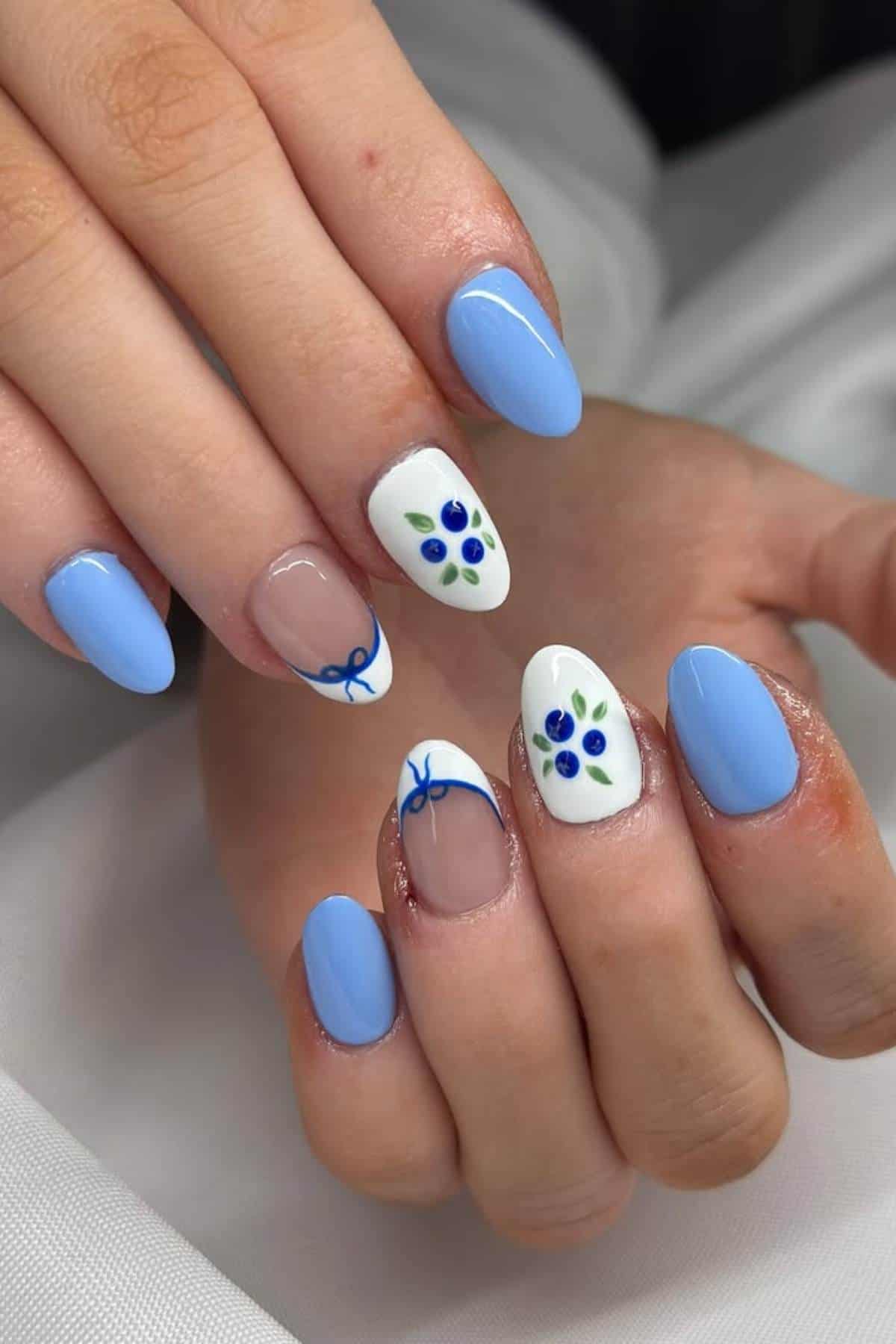 Hands with blue manicured nails; two nails feature white polish with blue and green floral designs, and two nails have small blue bow accents on a nude base.