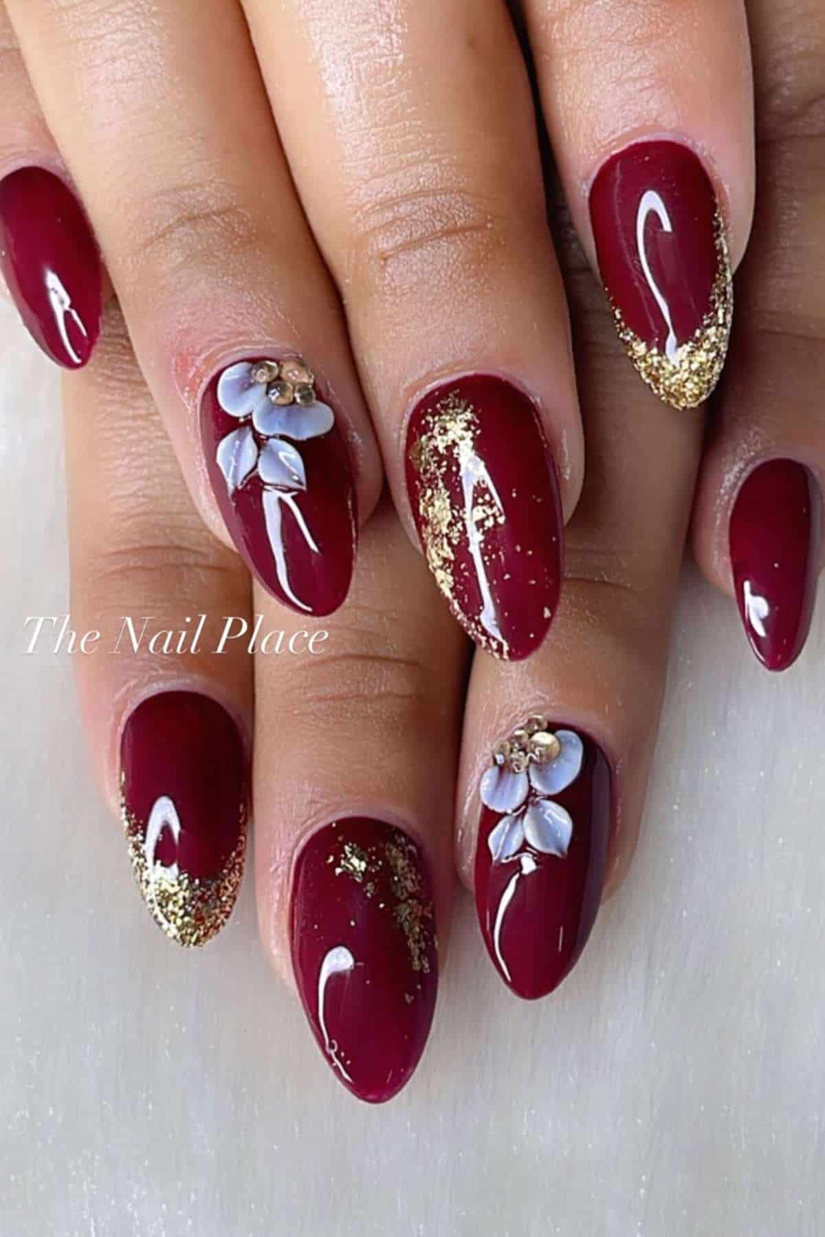 Hands with almond-shaped maroon gel nails, decorated with gold glitter and small 3D white flower accents on two fingers.