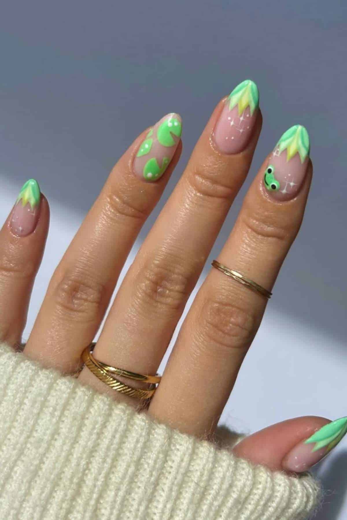 A hand with almond-shaped nails features green and yellow abstract nail art designs. The person is wearing gold rings and a cream-colored sweater.