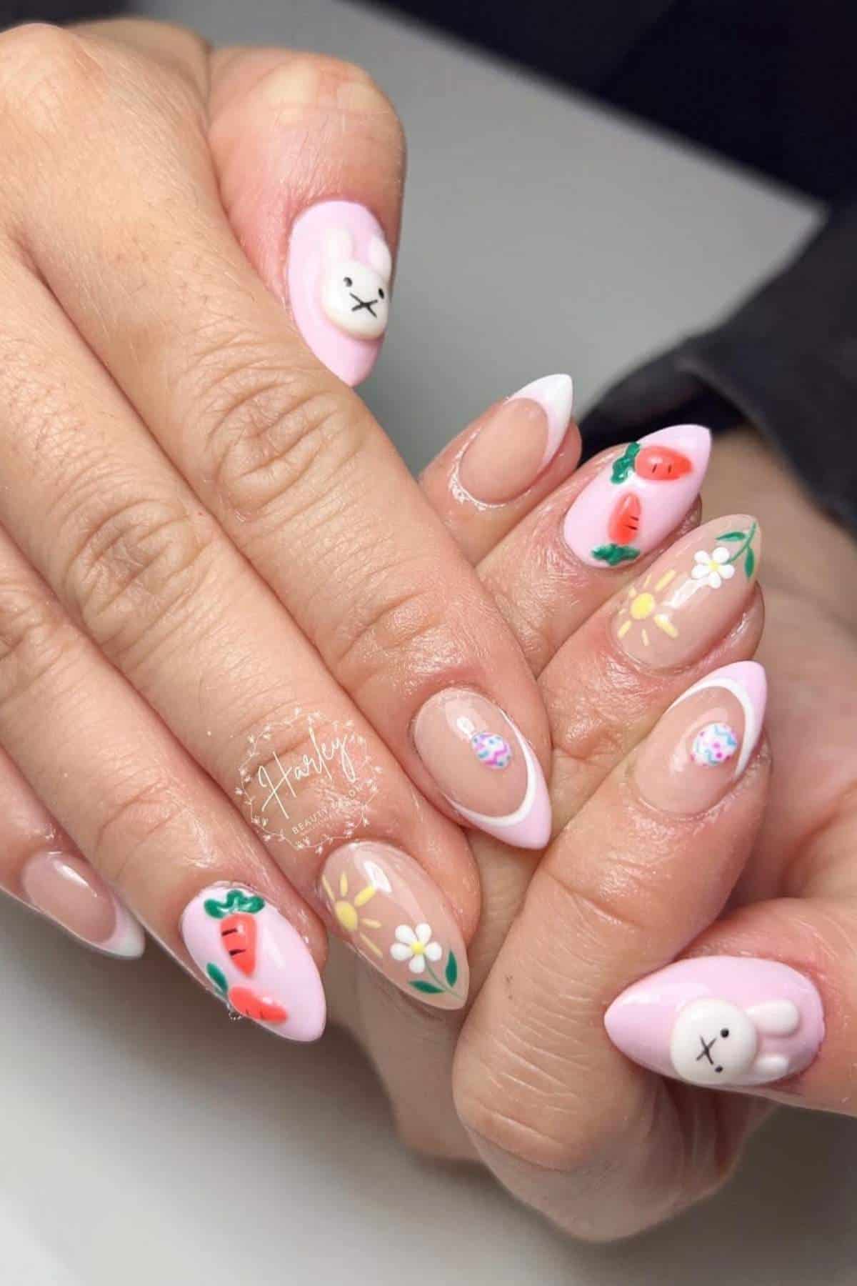 Hands with almond-shaped nails featuring pastel pink polish and designs of cartoon bunnies, carrots, strawberries, daisies, and small floral patterns.