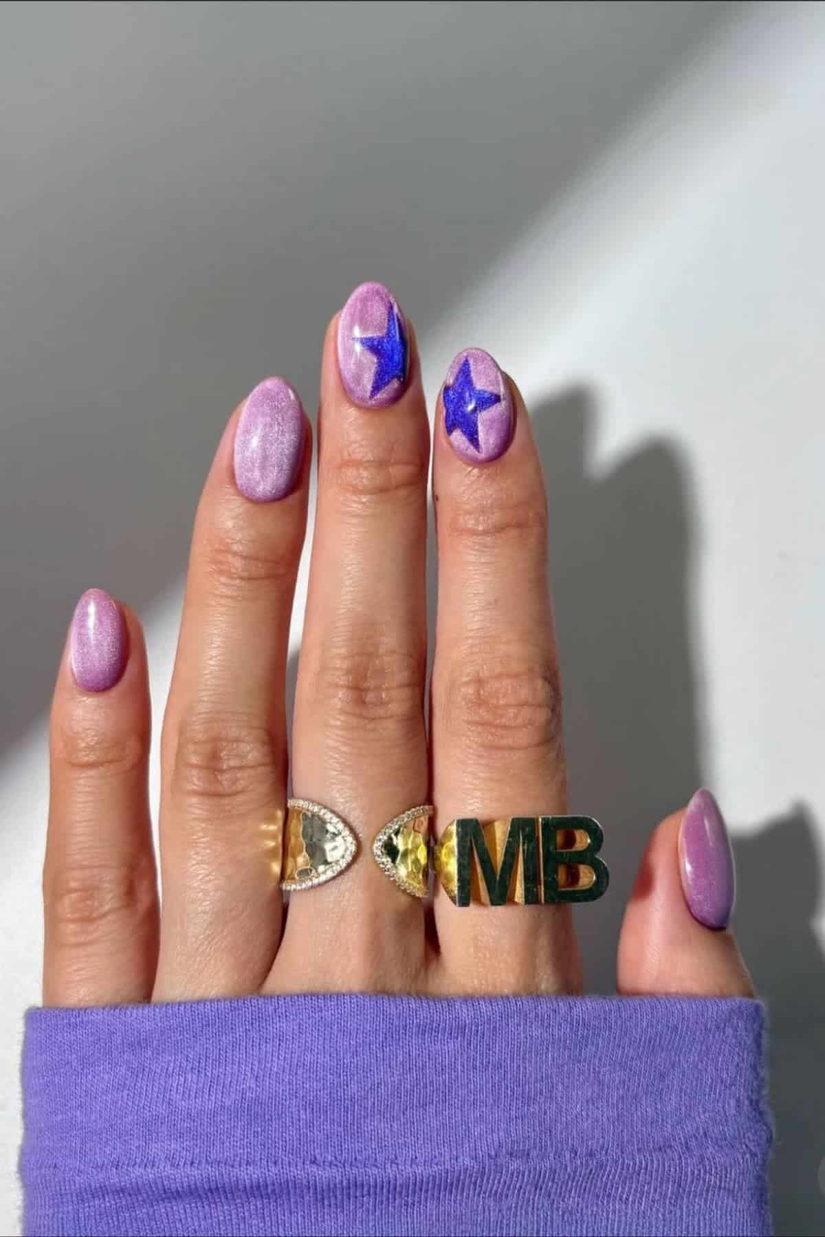 A hand with shimmering lavender nail polish and two blue star designs on the middle and ring fingernails, wearing gold rings, including one with the letters "MB," against a light background.