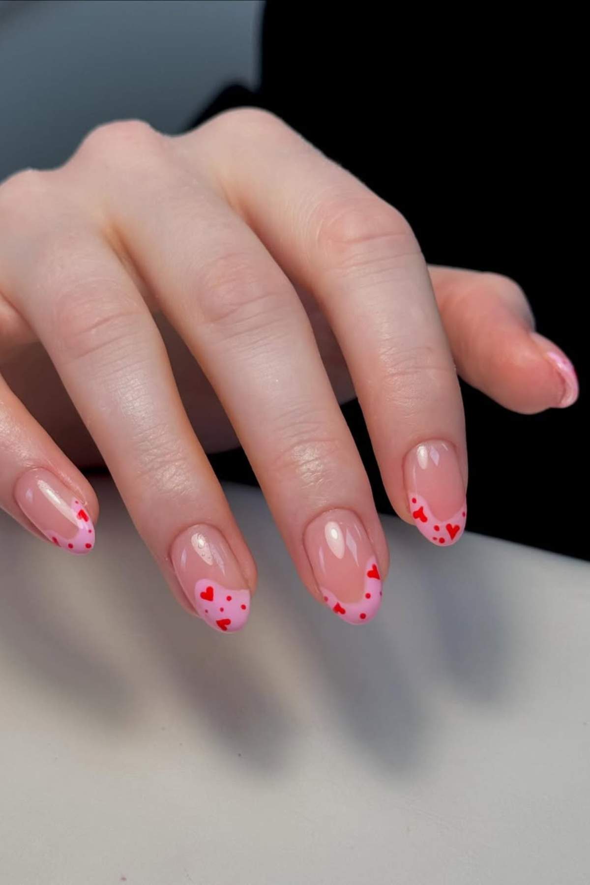 A hand with manicured nails featuring a nude base and red heart designs on the nail tips.
