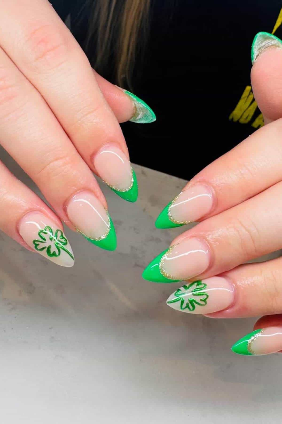 Hands with long almond-shaped nails featuring green French tips, gold accents, and shamrock designs on two accent nails.